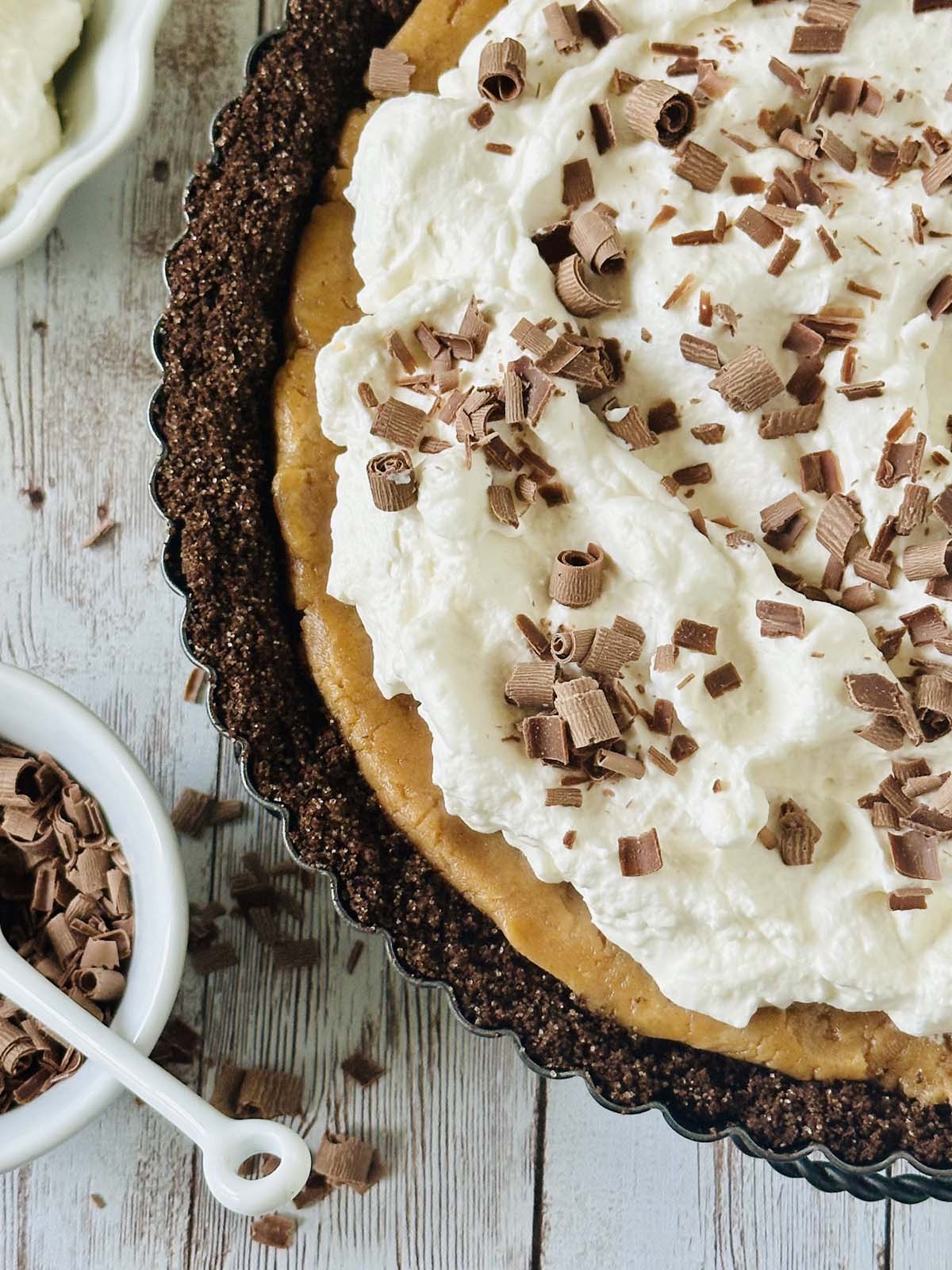A close-up of a peanut butter chocolate tart with a chocolate crust, topped with whipped cream and chocolate shavings, sits on a rustic wooden surface. A small bowl filled with more chocolate shavings is nearby.