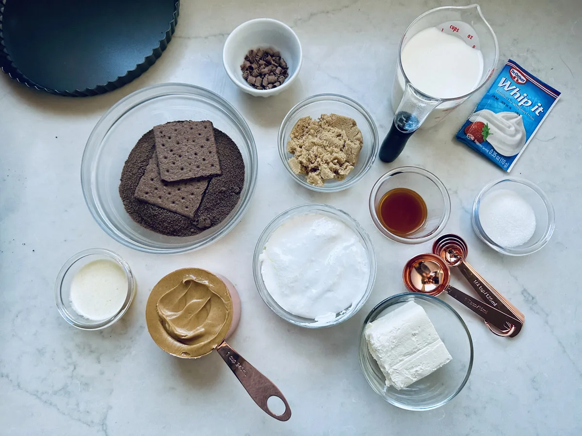 A variety of dessert ingredients for a peanut butter chocolate tart are on the countertop, including chocolate graham crackers, peanut butter, cream cheese, sugars, vanilla, whipped topping, chocolate chips, milk, and a tart pan.
