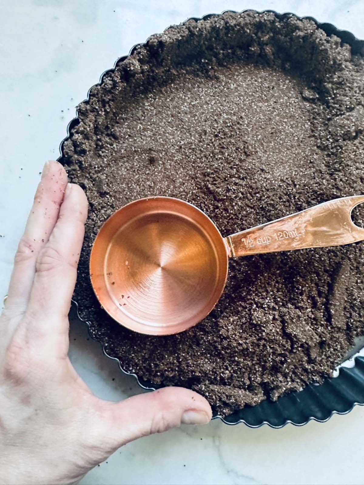 A hand presses a chocolate crumb crust into a round tart pan, using a copper measuring cup to help smooth and flatten the base for a decadent peanut butter chocolate tart.