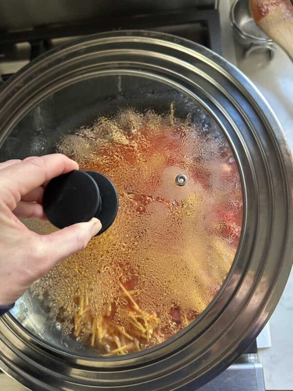 A hand lifts the lid of a slow cooker, revealing steam and condensation inside. Food, possibly noodles and sauce, is visible through the glass lid as it cooks.
