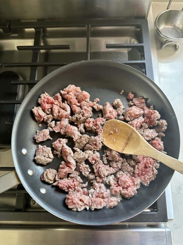 Ground beef is being browned in a black frying pan on a stovetop. A wooden spoon is stirring the meat, which is partially cooked, with some pieces still pink and others browned.