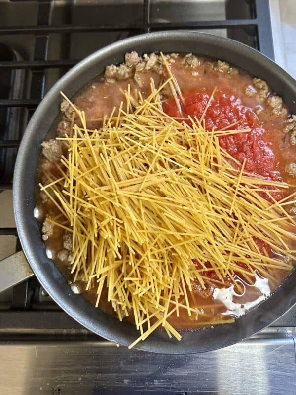A frying pan on a stovetop contains ground meat, tomato sauce, and uncooked spaghetti noodles, with the ingredients spread evenly and beginning to cook together.