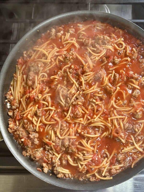 A skillet on a stovetop filled with cooked spaghetti, ground meat, and tomato sauce, with steam rising from the hot dish.