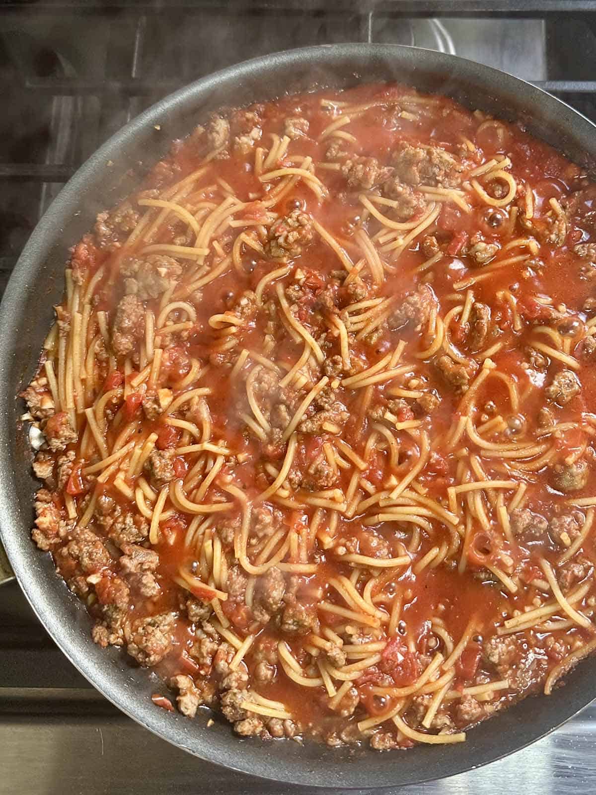 A skillet on a stovetop filled with cooked spaghetti, ground meat, and tomato sauce, with steam rising from the hot dish.