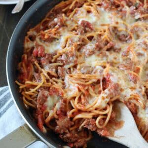 A skillet filled with baked spaghetti mixed with ground beef, tomato sauce, and melted cheese, with a wooden spoon resting inside. A plate with a serving of spaghetti and a fork is in the background.