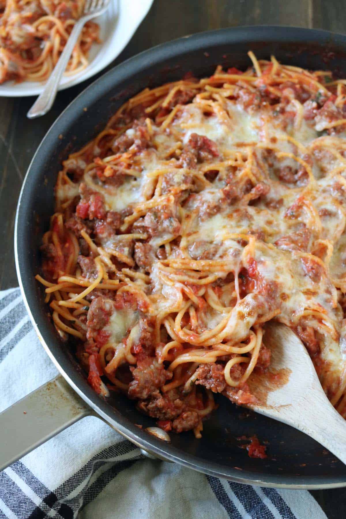 A skillet filled with baked spaghetti mixed with ground beef, tomato sauce, and melted cheese, with a wooden spoon resting inside. A plate with a serving of spaghetti and a fork is in the background.