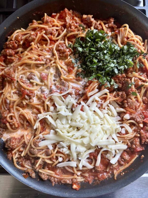 A skillet filled with spaghetti, ground meat in tomato sauce, topped with chopped fresh basil and shredded mozzarella cheese, before mixing.
