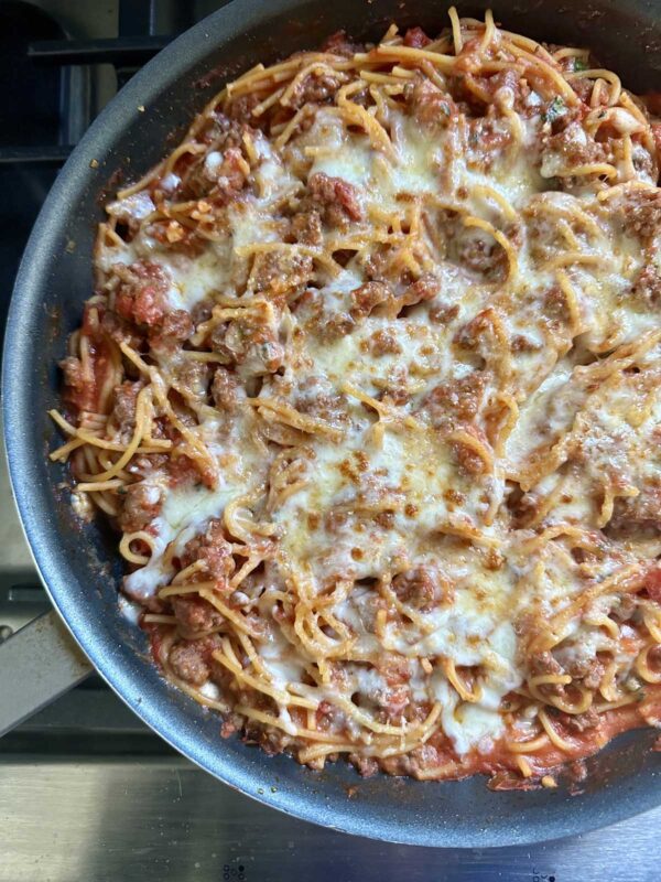 A pan filled with cooked spaghetti mixed with tomato sauce, ground meat, and topped with melted cheese, shown on a stovetop.