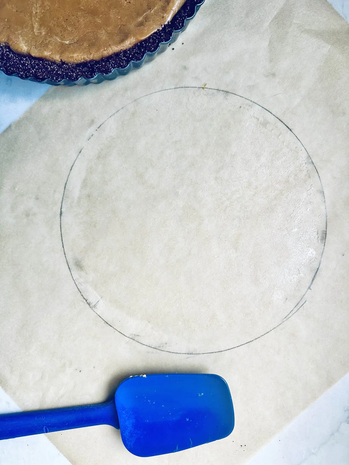 A sheet of parchment paper with a circle traced on it sits on a white surface. A blue spatula lies nearby, and part of a peanut butter chocolate tart in a pan is visible at the top of the image.