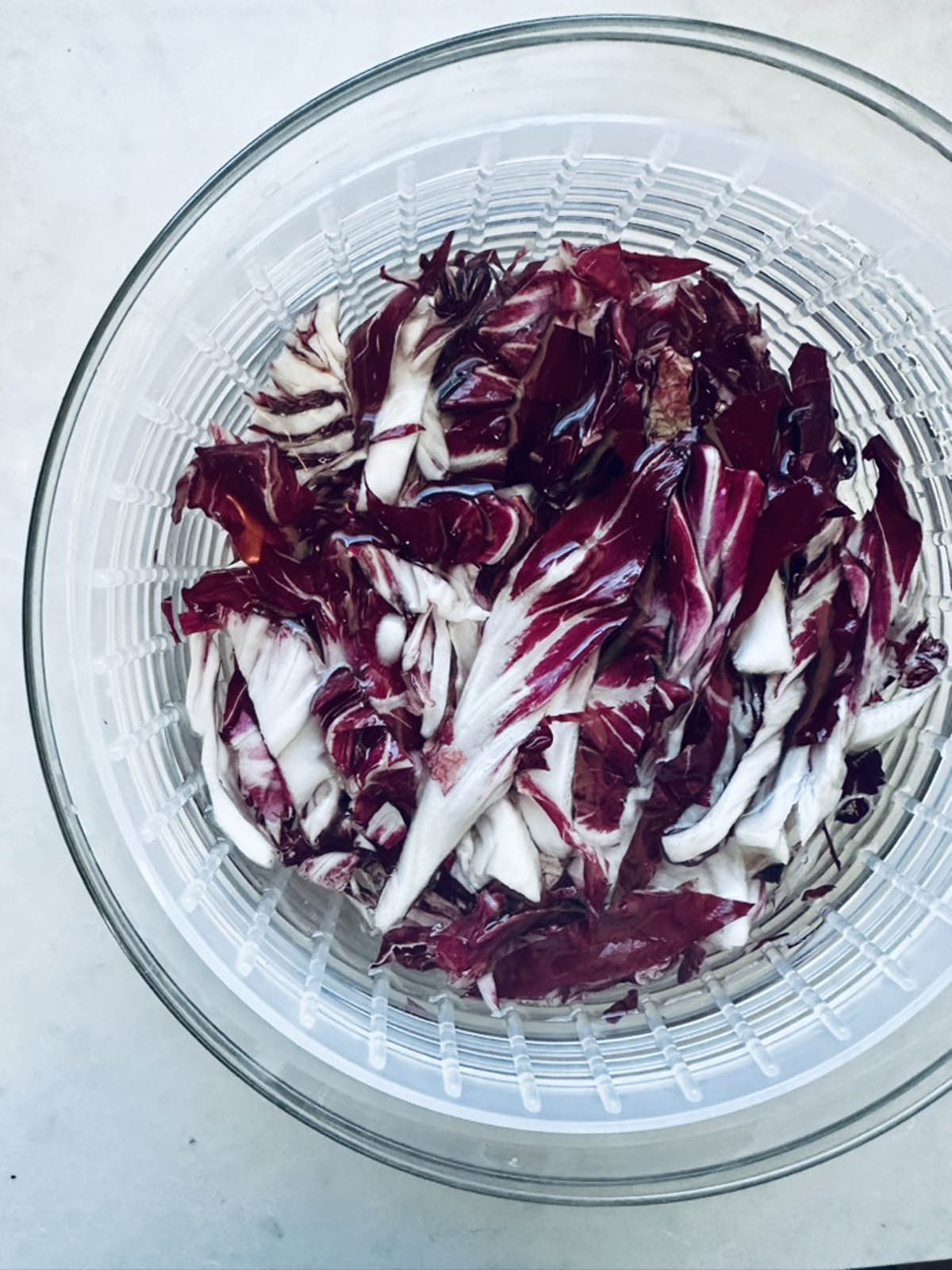 Shredded radicchio leaves in a clear salad spinner, viewed from above, showing their deep purple-red and white colors against a light background.