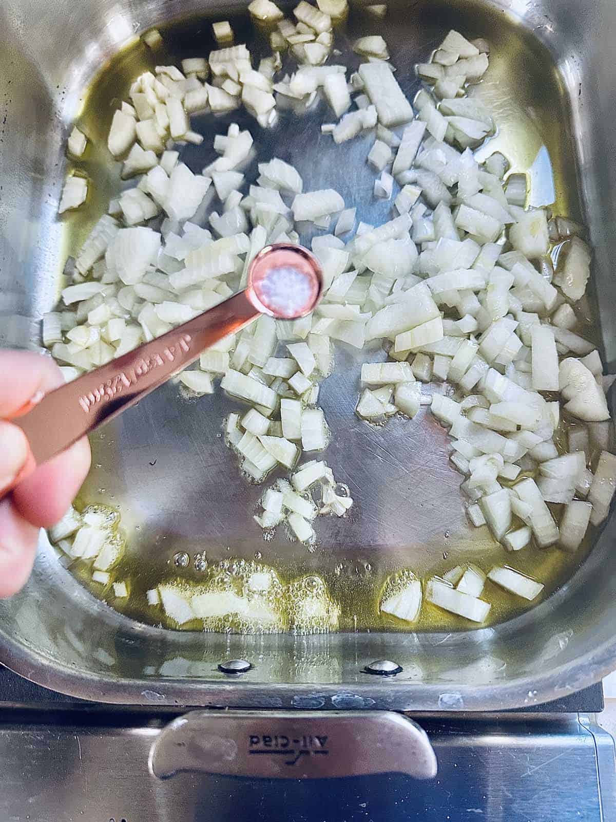 A hand holds a measuring spoon of salt above a pan with chopped onions saut&eacute;ing in oil on a stovetop.