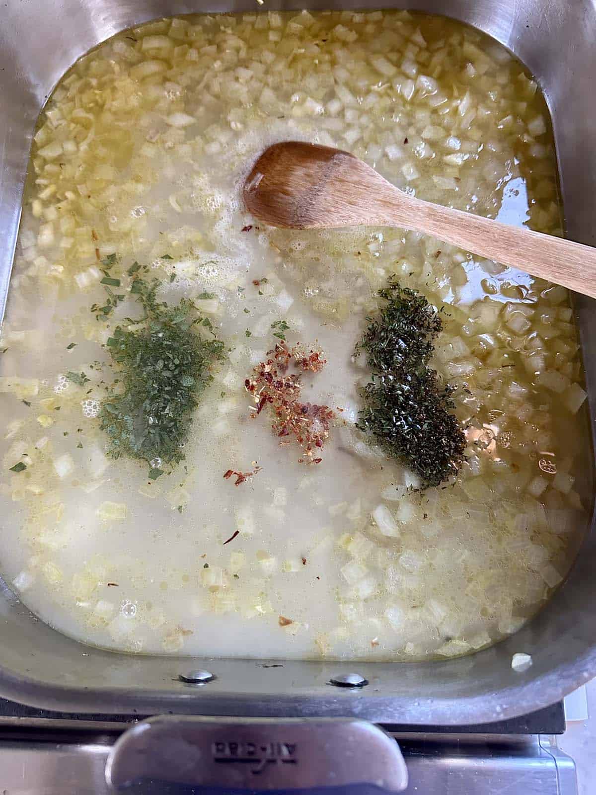 Chopped onions simmering in a pot of water with three piles of dried herbs and red pepper flakes, being stirred with a wooden spoon.