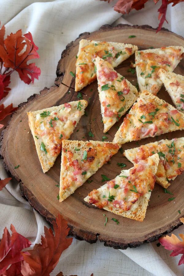 Slices of cheesy bread topped with bacon and herbs are arranged on a wooden serving board. Autumn leaves and a light cloth create a cozy, seasonal atmosphere around the board.