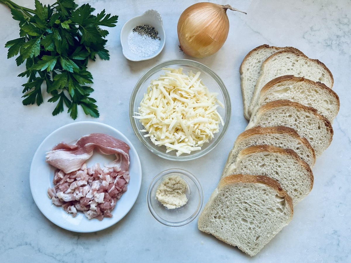 Slices of white bread, grated cheese, a whole onion, parsley, salt, pepper, raw bacon pieces, and grated Parmesan arranged neatly on a white surface.