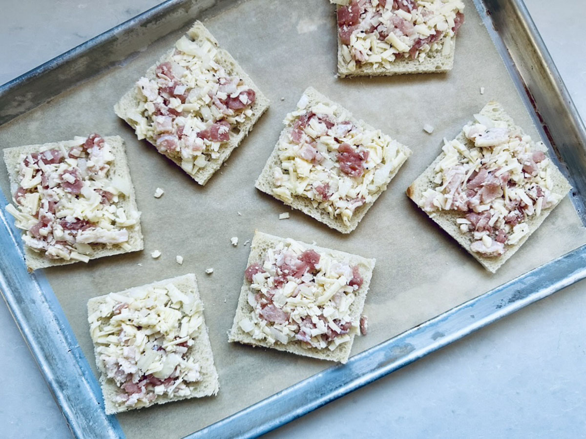 A baking tray lined with parchment paper holds seven square pieces of bread topped with grated cheese and small chunks of ham, arranged in a scattered layout.