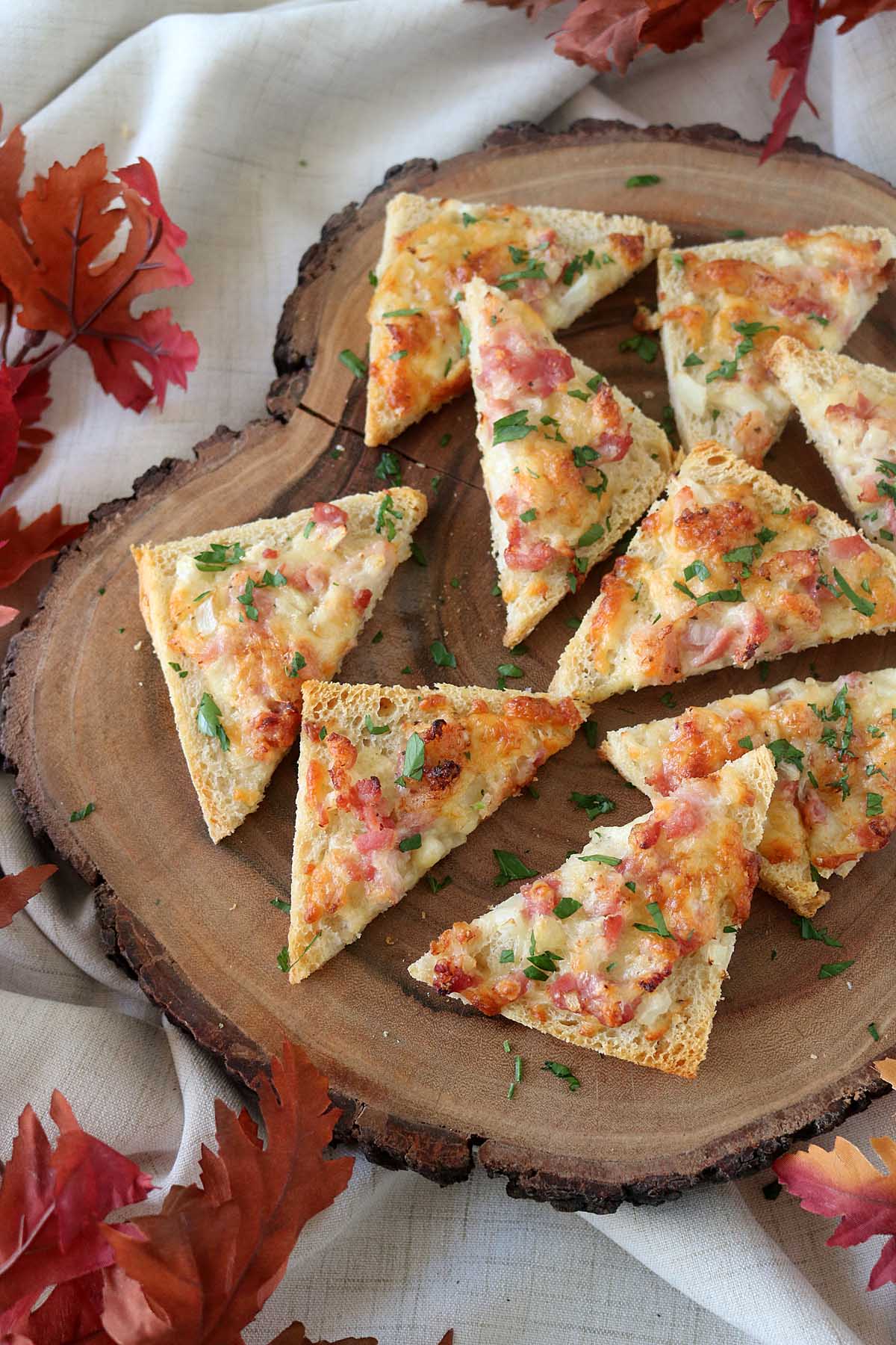 Slices of cheesy bread topped with bacon and herbs are arranged on a wooden serving board. Autumn leaves and a light cloth create a cozy, seasonal atmosphere around the board.