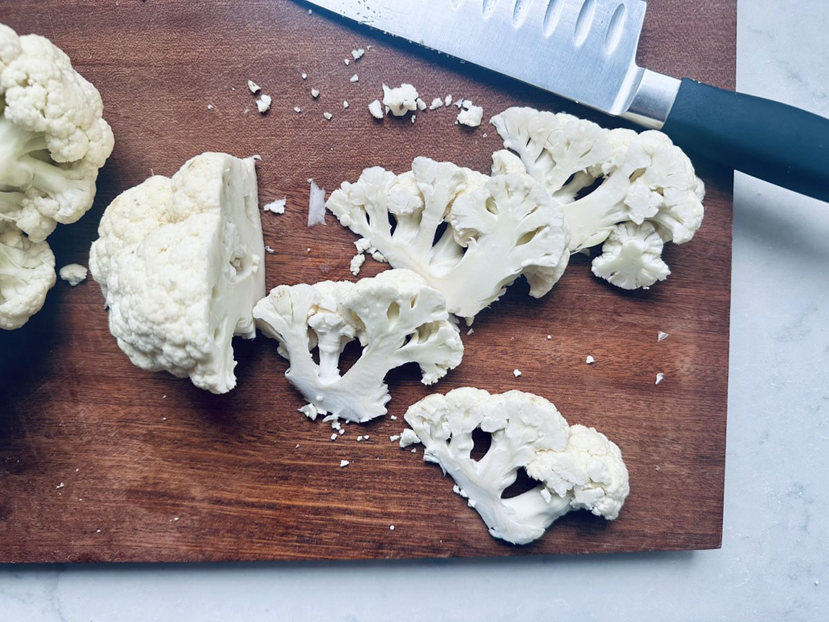 Several slices of raw cauliflower and a knife rest on a dark wooden cutting board, with small cauliflower pieces scattered around. Part of a whole cauliflower is visible on the left side of the board.