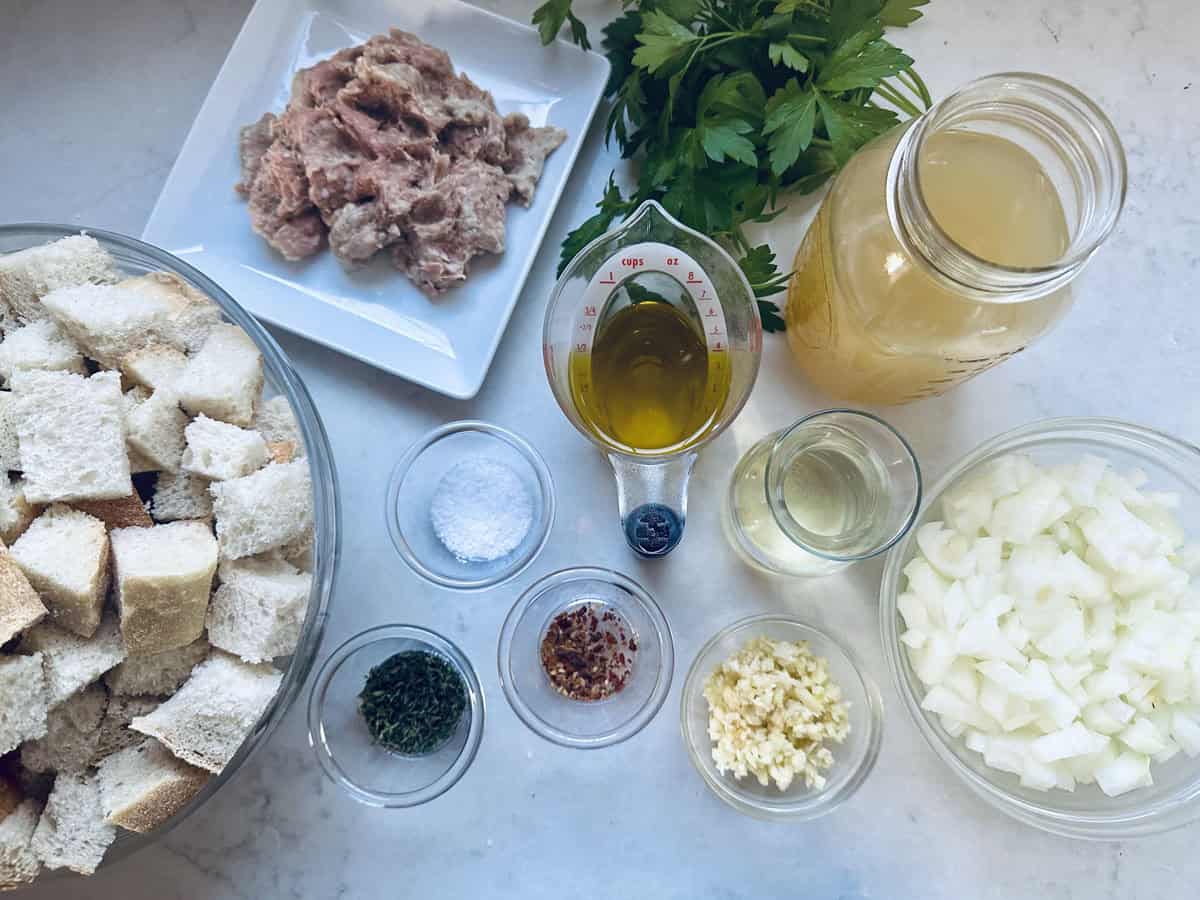 An overhead view of recipe ingredients: cubed bread, ground meat, chopped onion, minced garlic, broth, olive oil, parsley, salt, pepper, red pepper flakes, dried herbs, and white wine on a white surface.