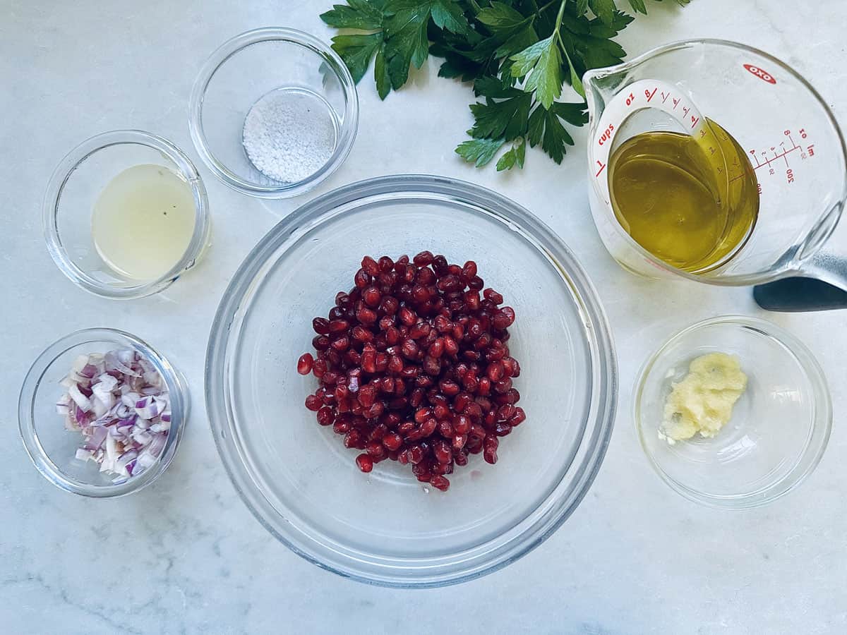 A glass bowl of pomegranate seeds is surrounded by bowls of chopped red onion, minced garlic, lemon juice, salt, olive oil, and fresh parsley on a white countertop.