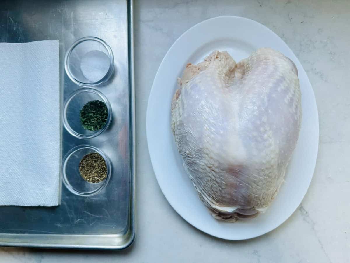 A raw turkey breast on a white plate sits beside a metal tray holding paper towels and three small bowls containing salt, dried parsley, and black pepper on a light countertop.