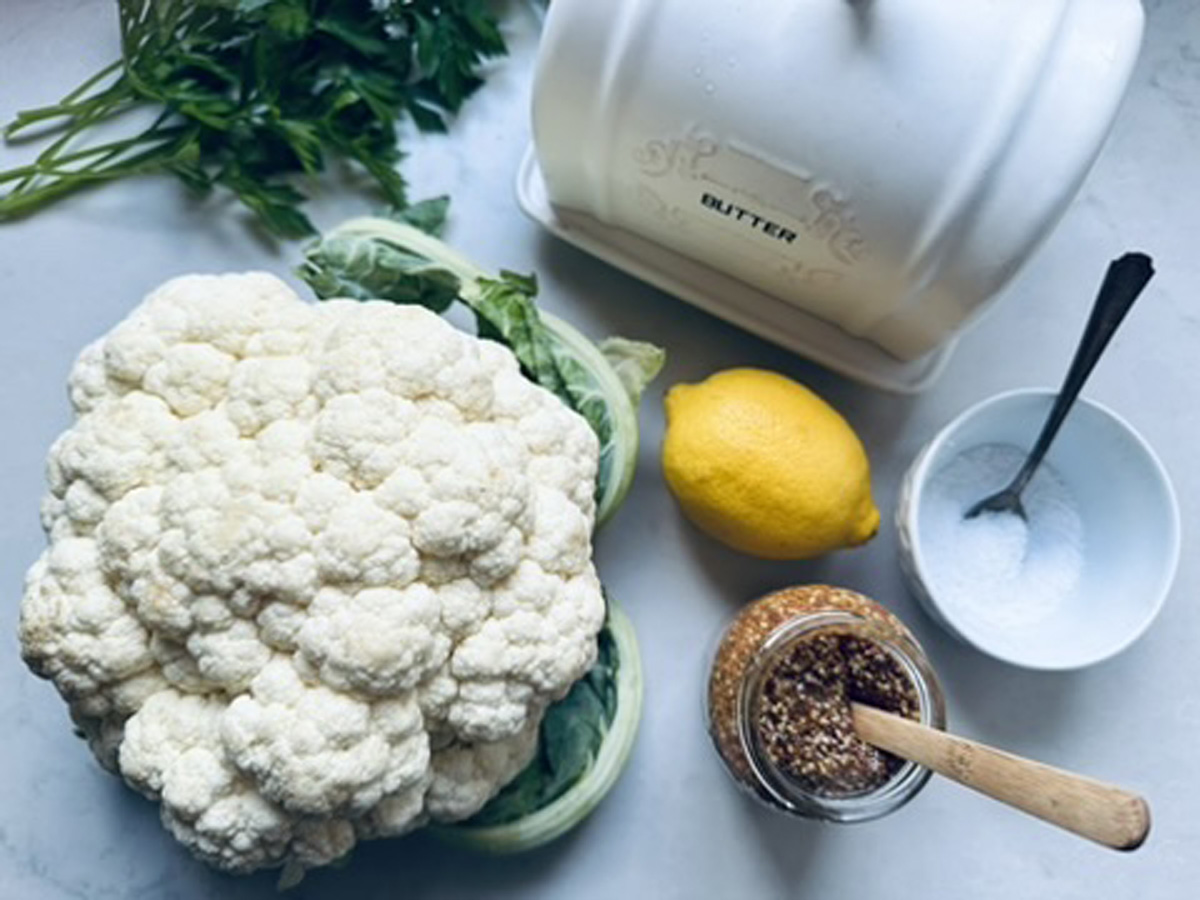 A head of cauliflower, a bunch of parsley, a lemon, a butter dish, a bowl of salt with a spoon, and a jar of grainy mustard are arranged on a white countertop.