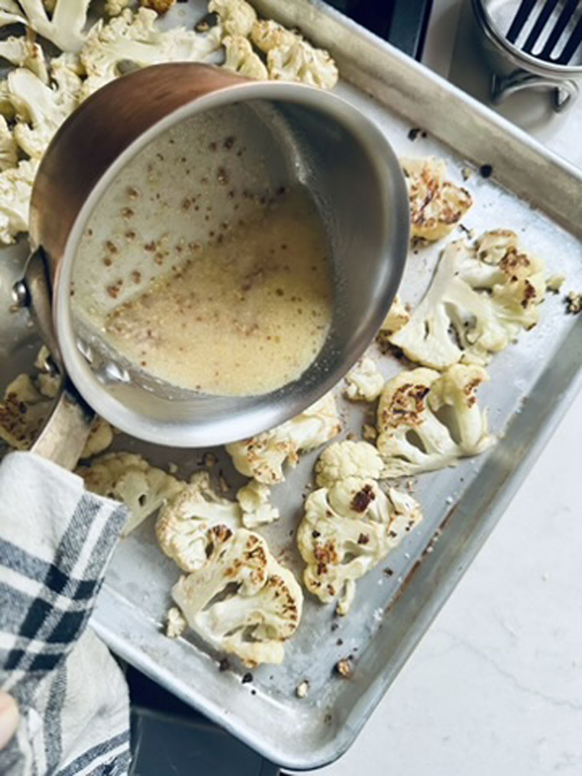 A saucepan of browned butter is being poured over roasted cauliflower florets on a baking sheet. A hand holding a striped kitchen towel grips the saucepan handle.
