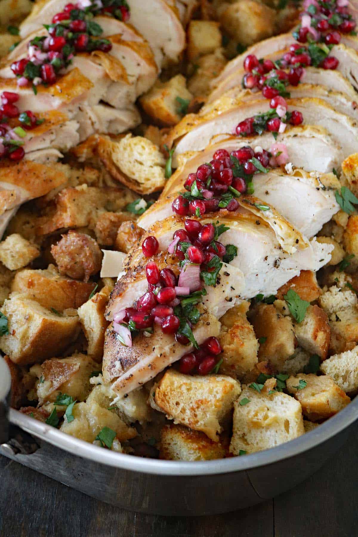 Sliced roasted turkey breast topped with pomegranate seeds and herbs, served over a bed of golden, cubed bread stuffing in a metal pan.