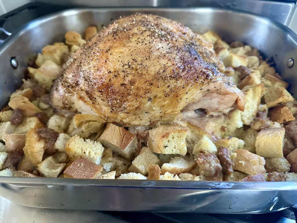 A roasted turkey breast sits atop a bed of cubed bread stuffing in a metal roasting pan, with the bread cubes browned and edges crisp from baking.