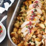 A roasting pan filled with cubed bread stuffing, topped with sliced roasted turkey breast garnished with herbs and pomegranate seeds. A spoon and napkins are on the side.