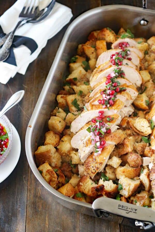A roasting pan filled with cubed bread stuffing, topped with sliced roasted turkey breast garnished with herbs and pomegranate seeds. A spoon and napkins are on the side.