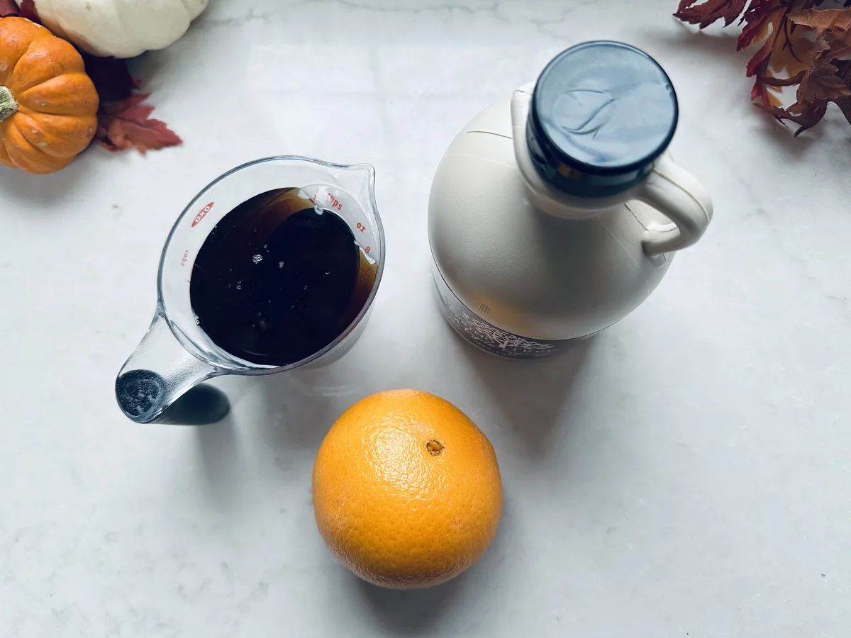 A measuring cup filled with dark liquid, a bottle of maple syrup, and a whole orange are arranged on a white surface with small pumpkins and autumn leaves in the corners.
