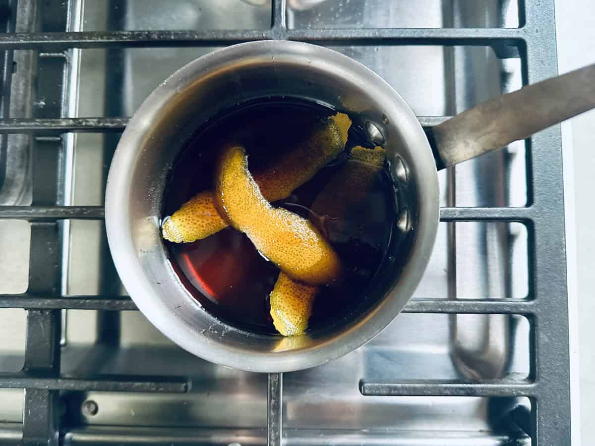 A metal pot with dark liquid and several orange peels sits on a stovetop burner, viewed from above.