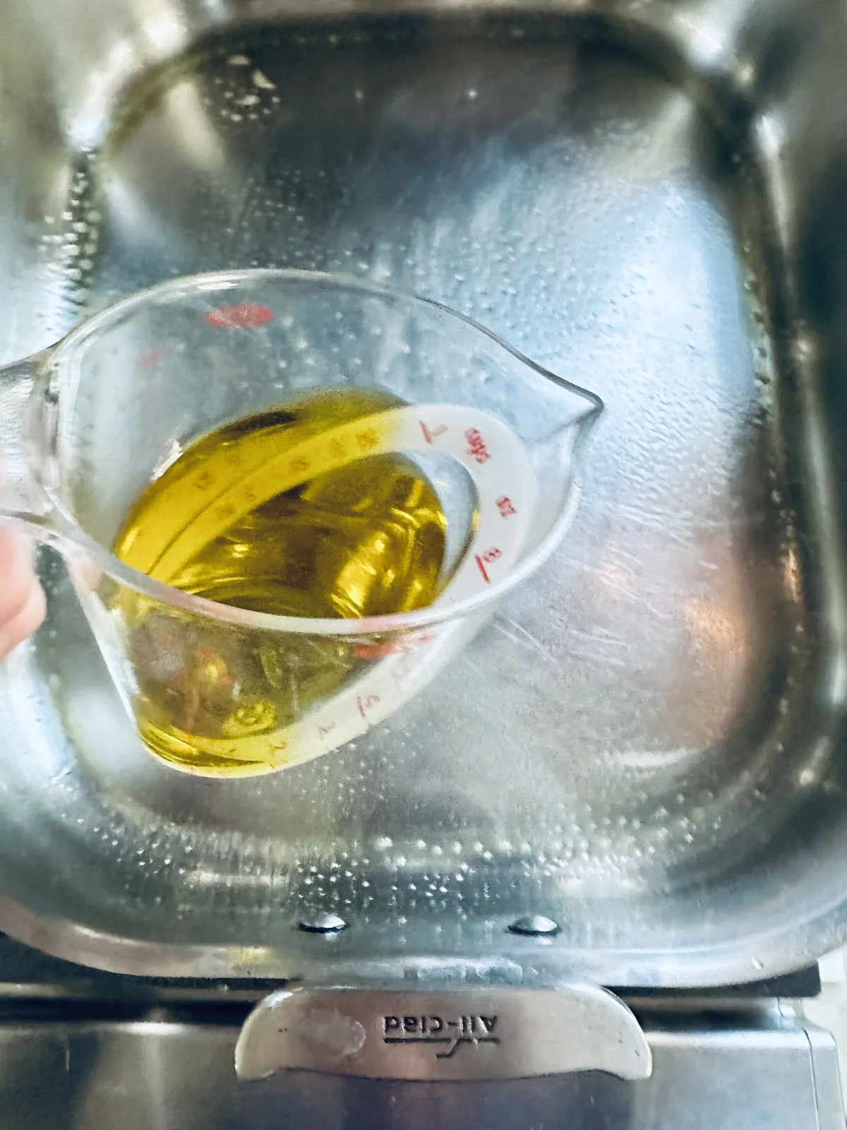 A hand holds a glass measuring cup filled with yellow oil above a metal All-Clad slow cooker insert.