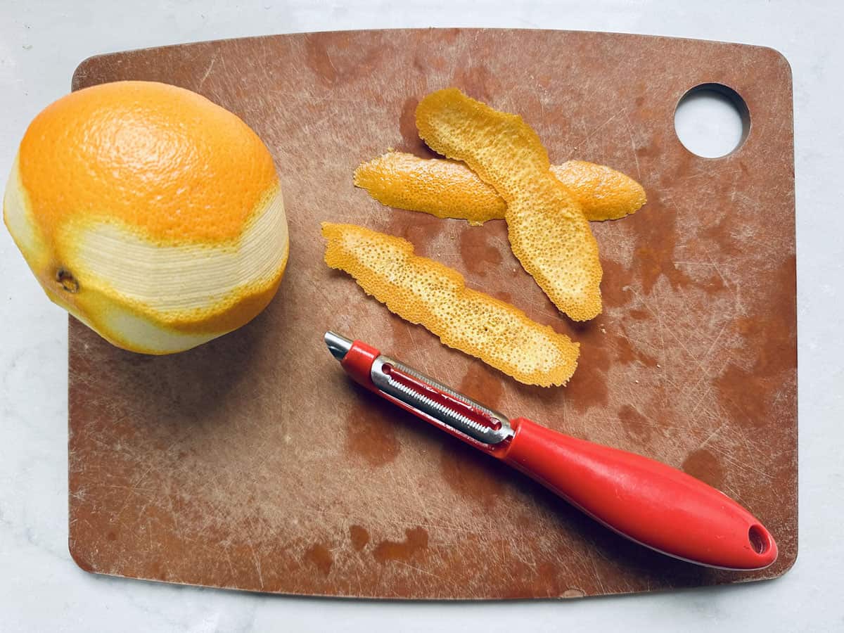 A partially peeled orange, three strips of orange peel, and a red-handled vegetable peeler are on a brown cutting board with a hole in the corner.