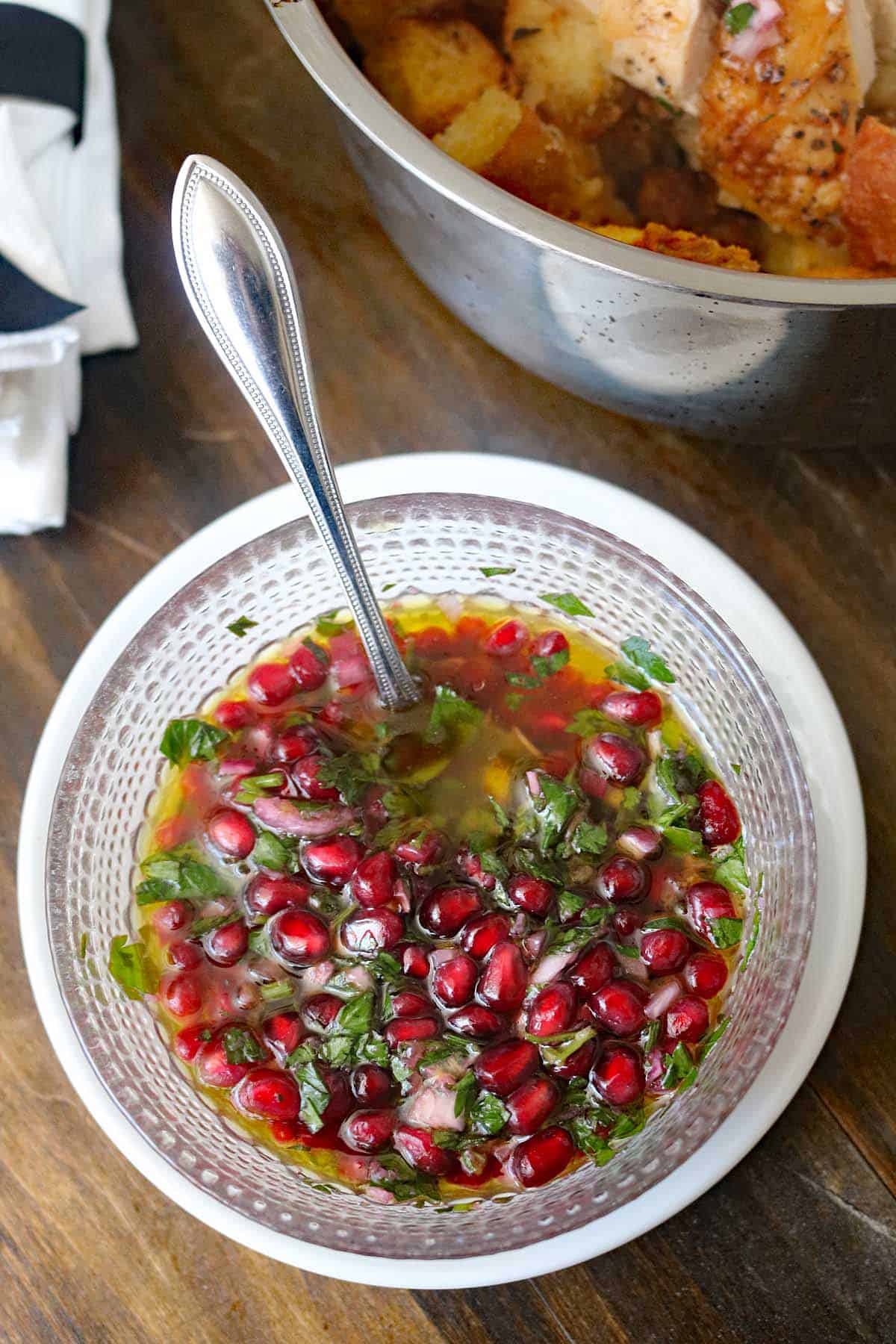A clear bowl filled with a vibrant sauce containing pomegranate seeds, chopped herbs, and olive oil, with a spoon inside; a pot of food is partially visible in the background.