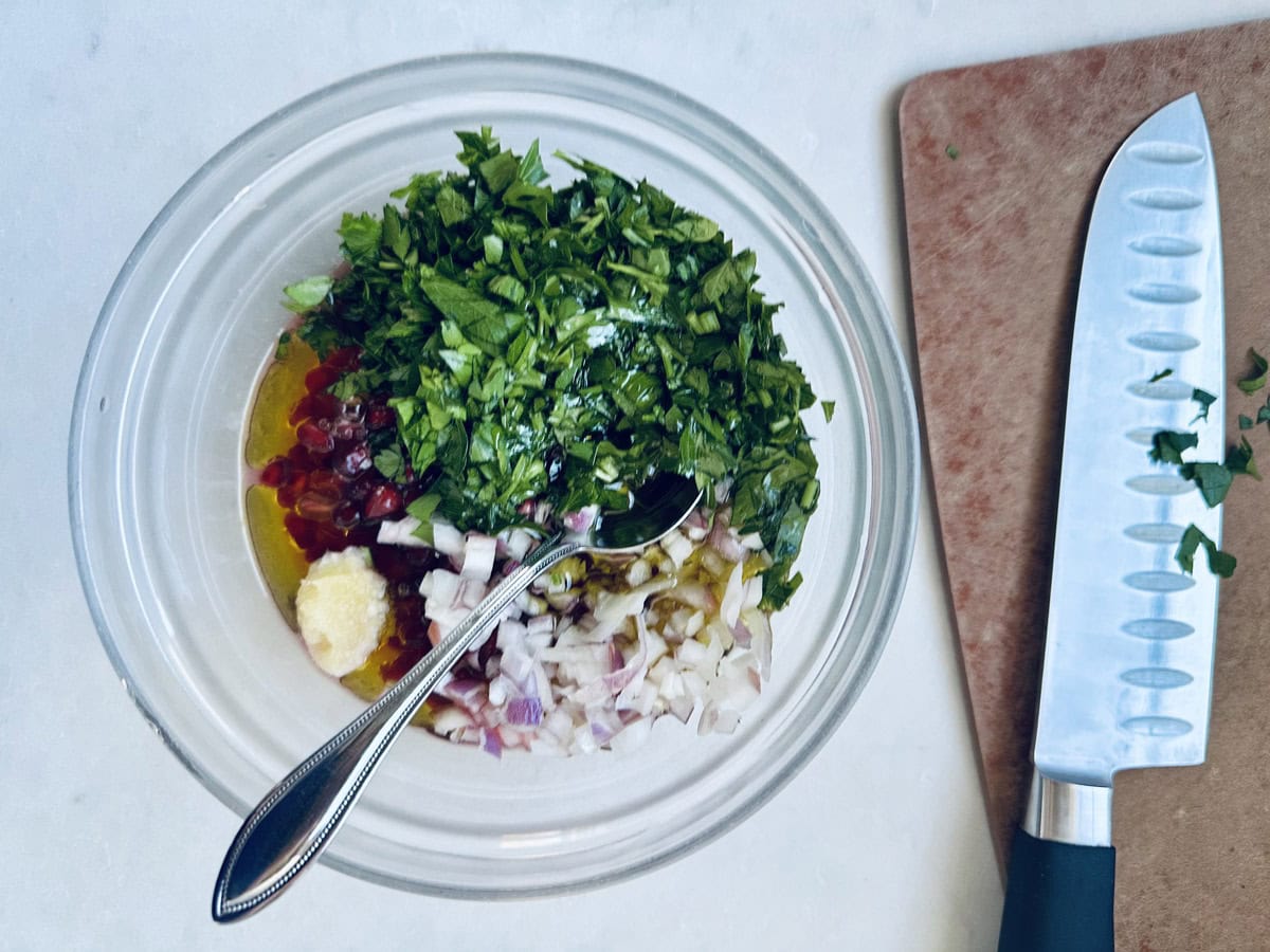 A glass bowl contains chopped parsley, diced onions, minced garlic, olive oil, and chopped red chili with a spoon inside. Next to it is a knife on a cutting board with herb pieces on both.