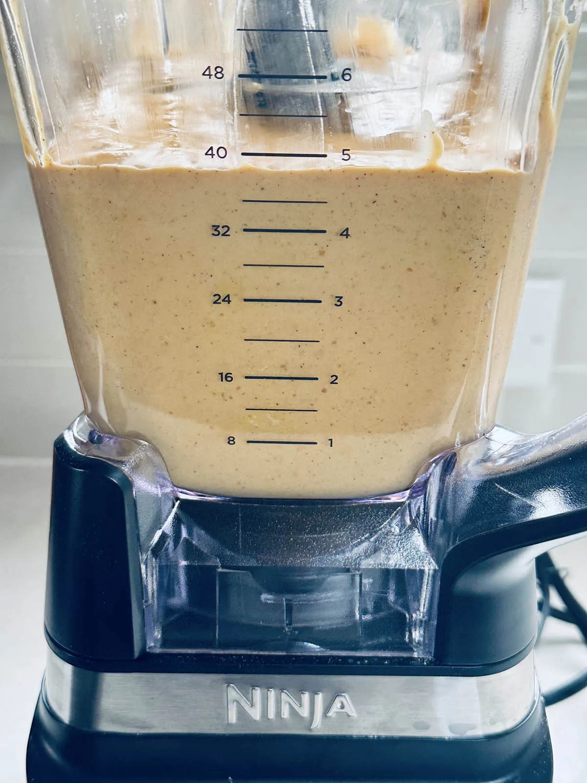 A close-up of a Ninja blender filled with a creamy, tan-colored mixture. The blender&rsquo;s measurement markings are visible on the container, and the appliance is sitting on a kitchen counter.