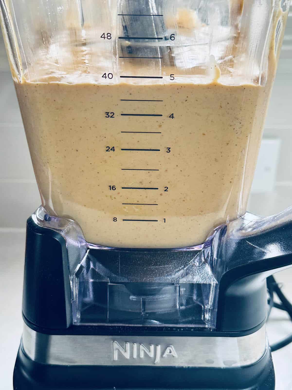 A close-up of a Ninja blender filled with a creamy, tan-colored mixture. The blender’s measurement markings are visible on the container, and the appliance is sitting on a kitchen counter.