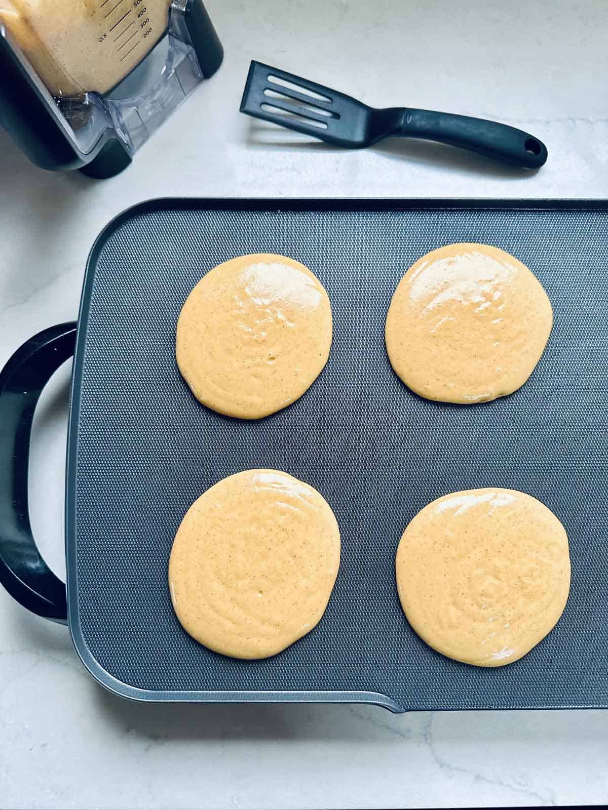 Four round pancake batters cooking on a nonstick griddle, with a black spatula and a blender of extra batter nearby on a light-colored countertop.