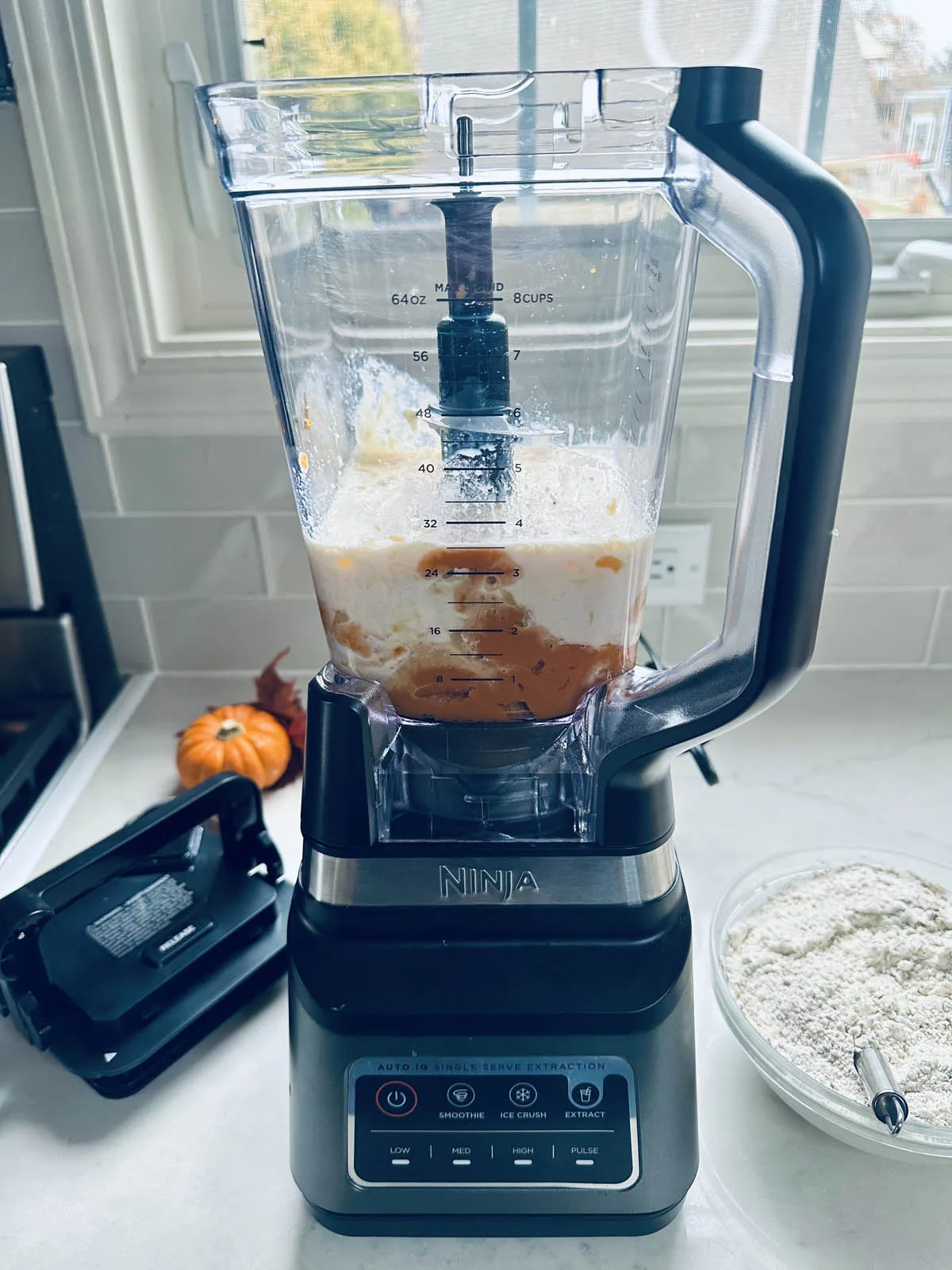 A Ninja blender filled with milk and orange ingredients sits on a kitchen counter beside a bowl of flour, a spoon, and a small pumpkin. The blender lid is off and placed to the side.