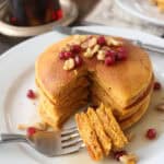 A stack of pumpkin pancakes topped with walnuts and pomegranate seeds sits on a white plate. A fork holds a cut bite, and syrup is drizzled over the pancakes. A syrup jar is in the background.