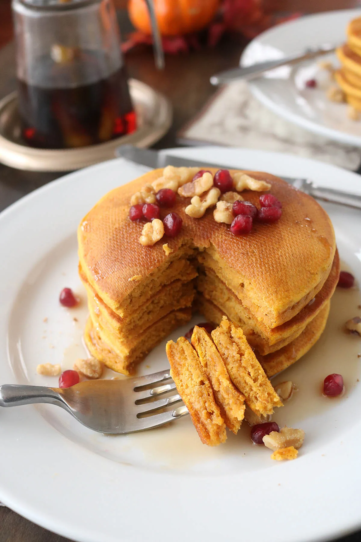 A stack of pumpkin pancakes topped with walnuts and pomegranate seeds sits on a white plate. A fork holds a cut bite, and syrup is drizzled over the pancakes. A syrup jar is in the background.