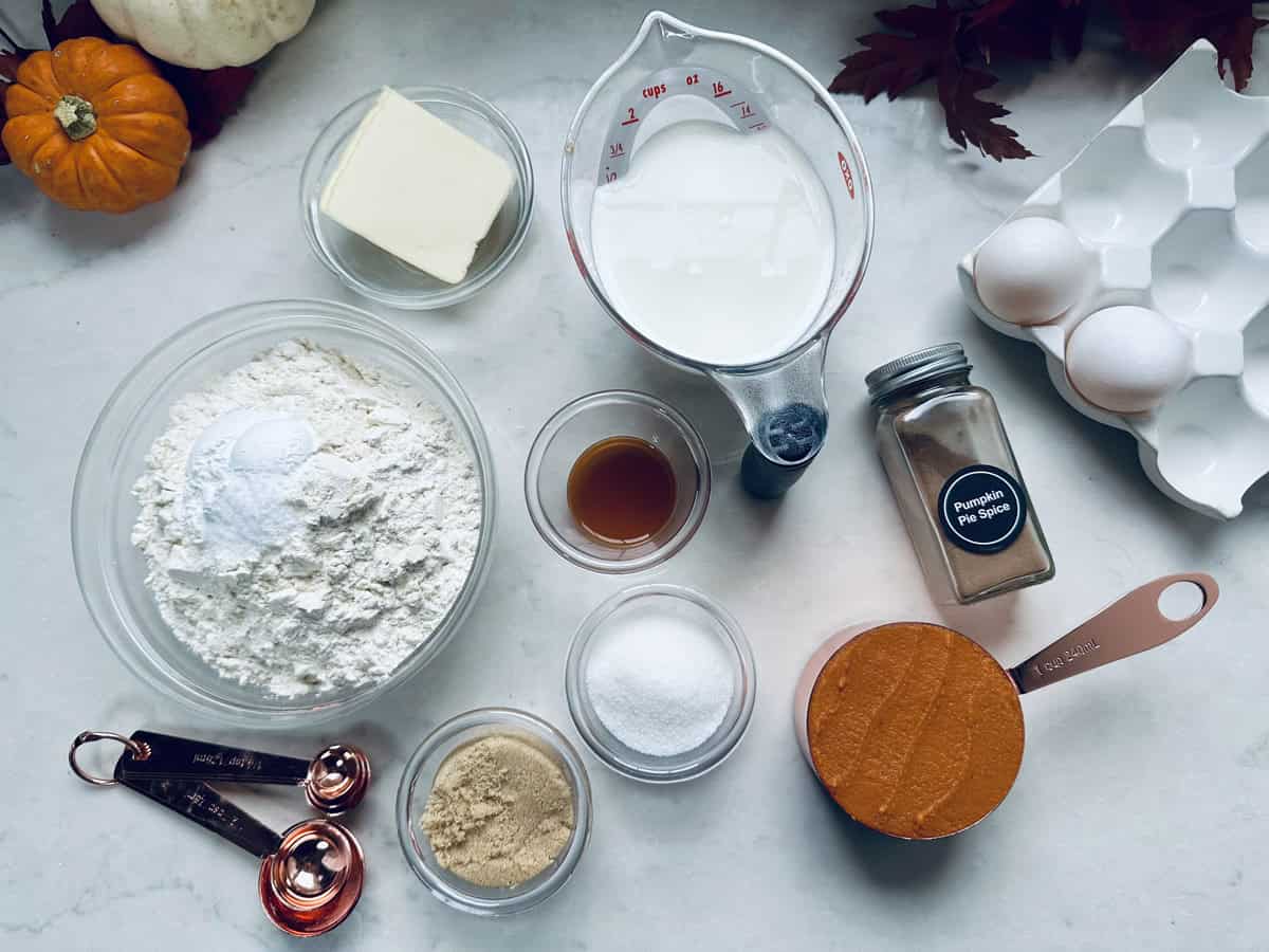 A top-down view of baking ingredients on a white surface, including flour, butter, milk, eggs, pumpkin puree, sugar, brown sugar, vanilla extract, pumpkin pie spice, and measuring spoons. Small pumpkins and leaves are nearby.