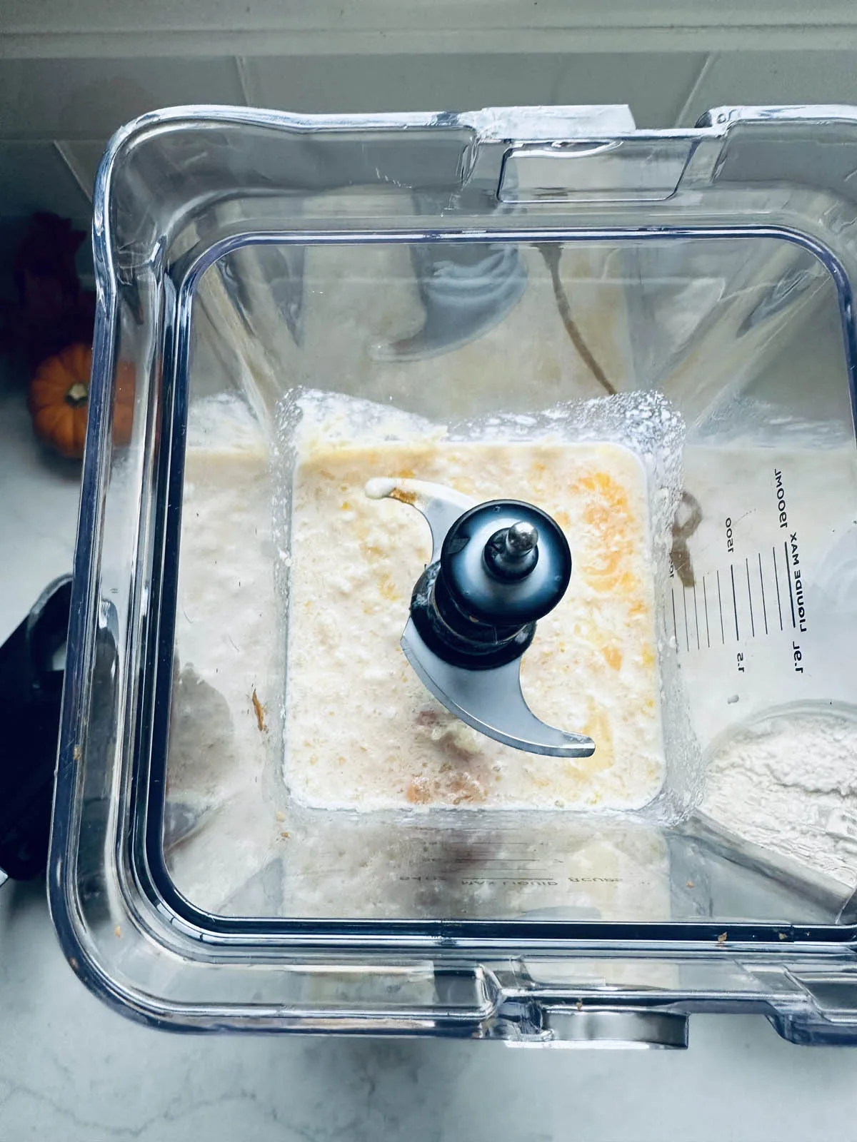 A top-down view of a blender containing a mixture of wet and dry ingredients, including visible lumps and liquids, ready to be blended. The blender sits on a white countertop.