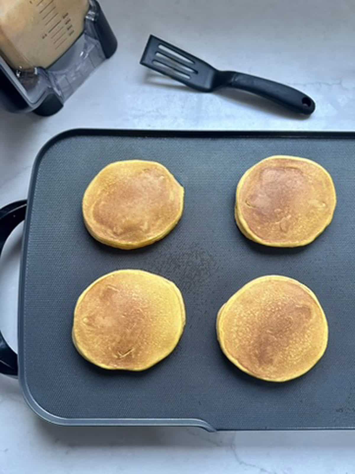 Four golden-brown pancakes cooking on a nonstick electric griddle, with a black spatula and a blender nearby on a white countertop.