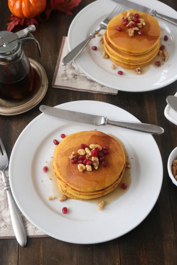 Two plates with stacks of pancakes topped with pomegranate seeds and walnuts sit on a wooden table. Nearby are a syrup dispenser, cutlery, napkins, and autumn-colored decor.
