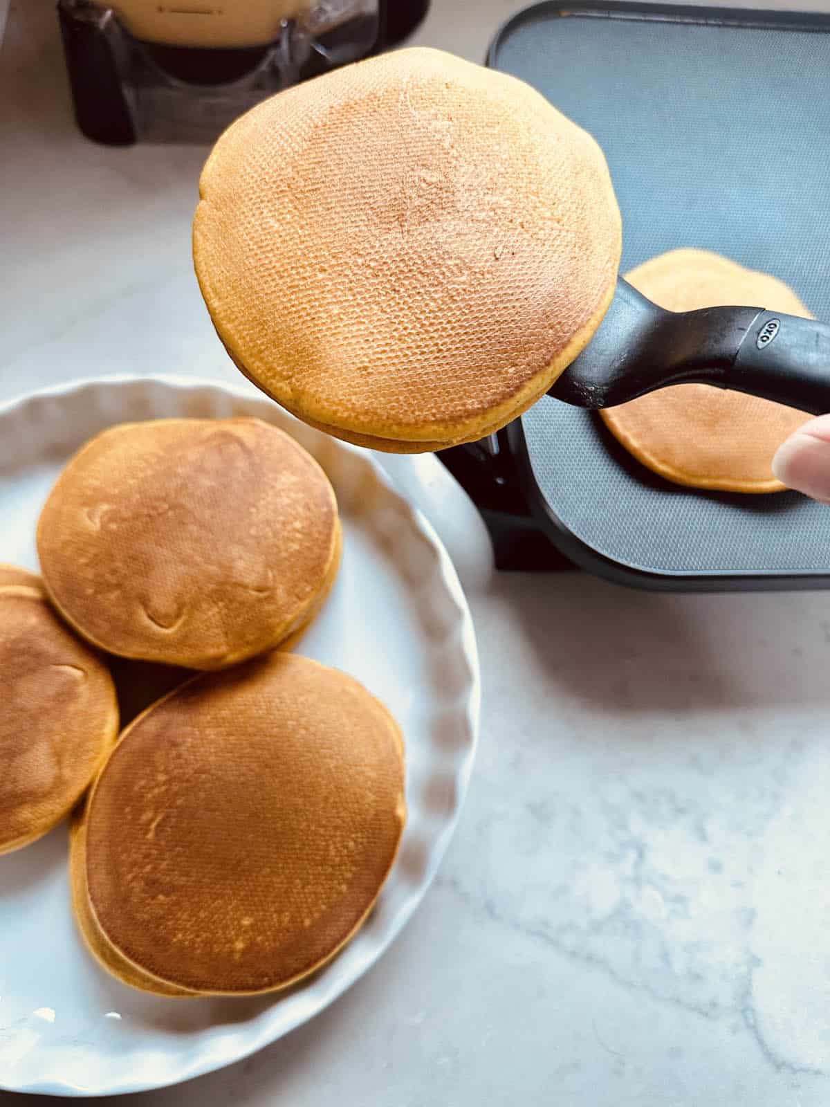 A hand holds a spatula lifting a golden brown pancake from a griddle, with more pancakes on a white plate nearby.