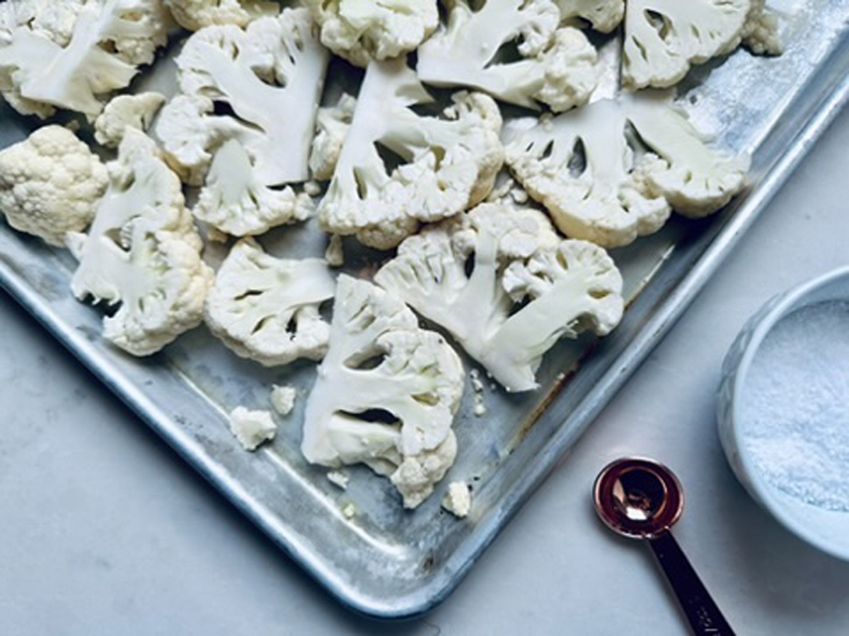 Sliced cauliflower florets spread on a metal baking sheet, next to a measuring spoon and a small bowl filled with salt on a white surface.