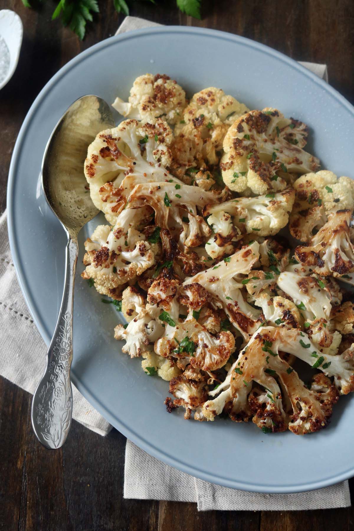 A blue plate with roasted cauliflower florets garnished with chopped herbs, served with a silver spoon, placed on a cloth napkin on a wooden table.