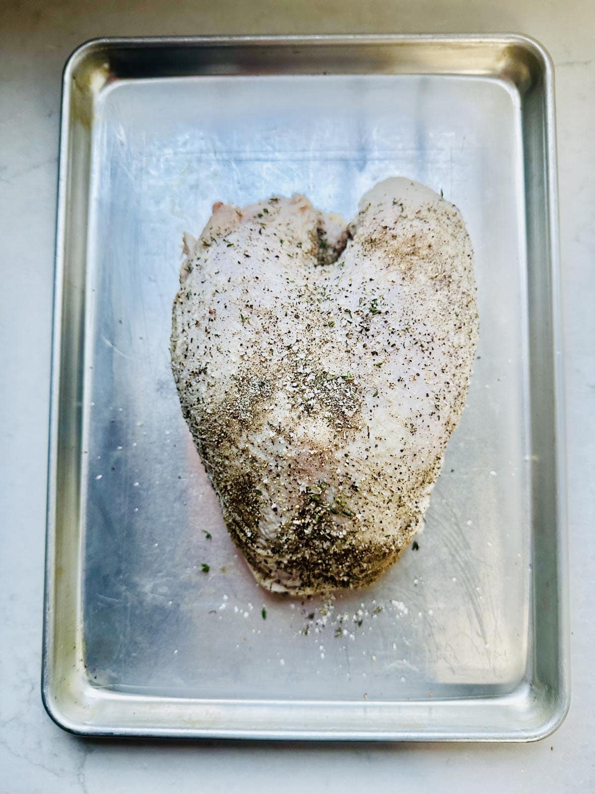 A raw, seasoned chicken breast in the shape of a heart sits on a metal baking tray against a light background.
